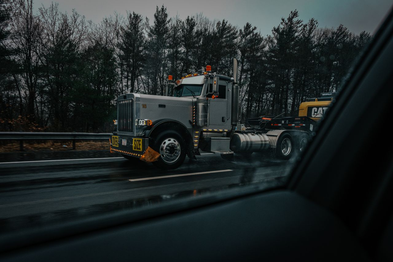 A large truck with an oversize load on a rainy highway, captured through a car window with dark, moody tones.